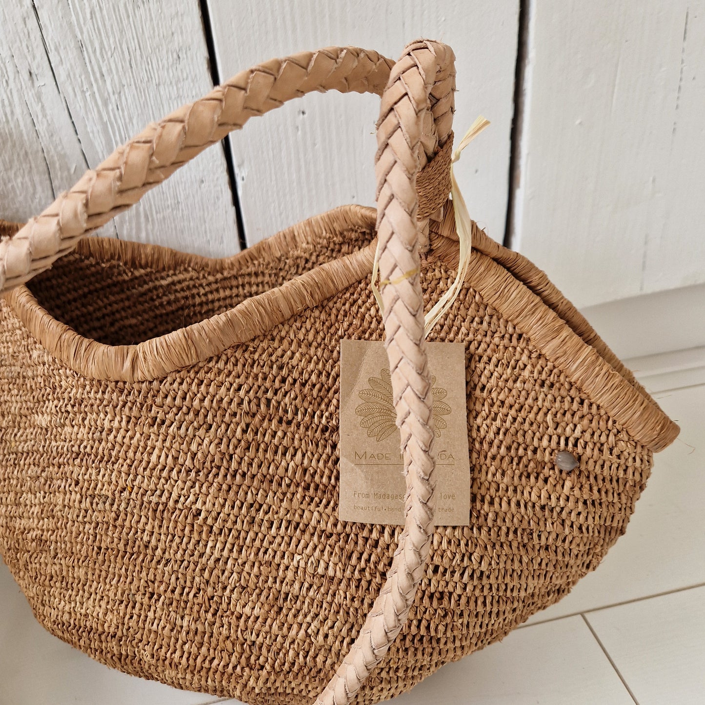 Detail van de Aina M Bag Light Brown met gevlochten lederen hengsel en by Bliss-label, rustend op witte vloer