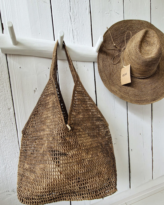 Raffia tas Ombinisoa  in de kleur tea van Made in Mada samen met de Elya hoed van Made in Mada aan een witte kaptstok
