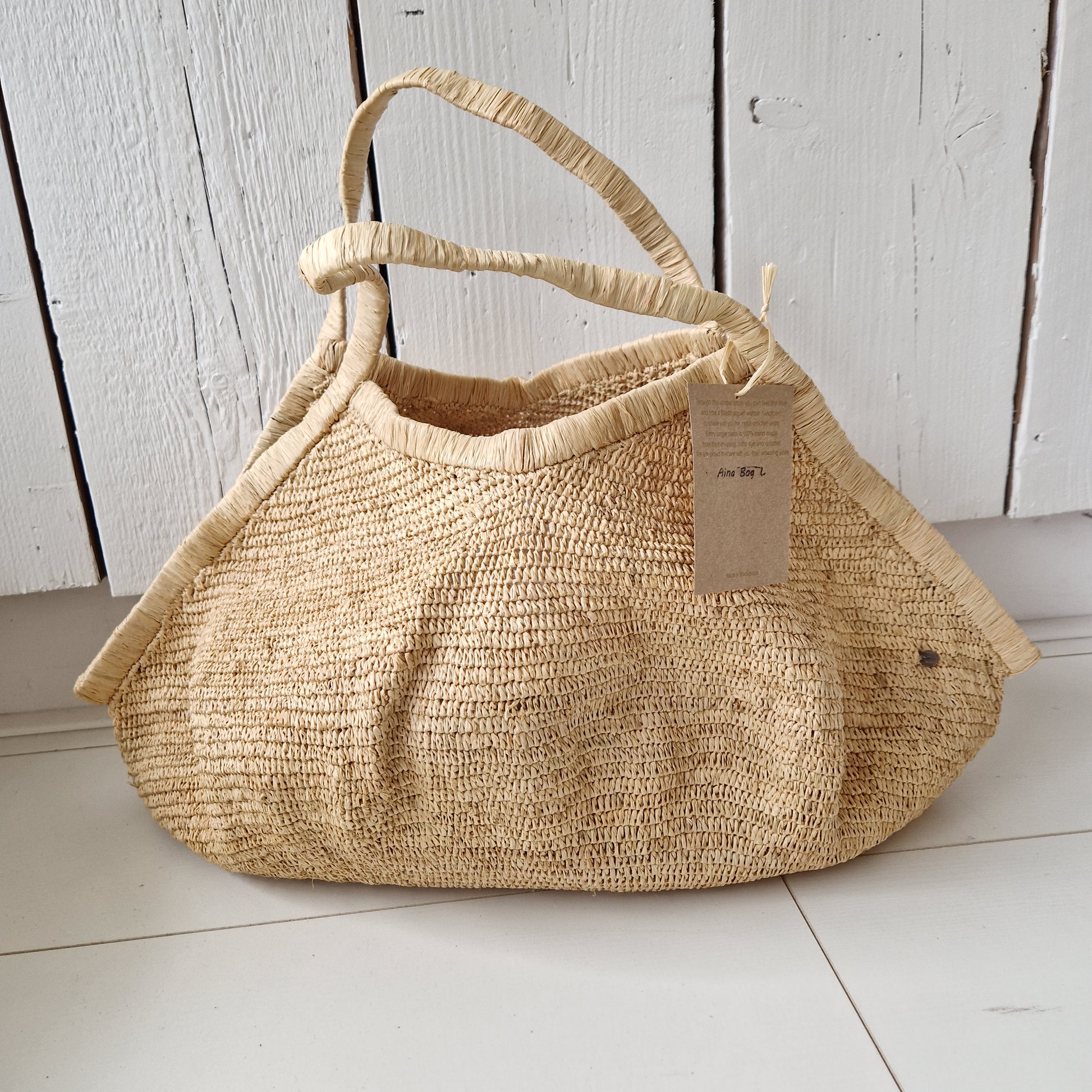 Aina Bag L Naturel van Made in Mada rustend op witte vloer, handgemaakte raffiatas met organische vorm en lichte uitstraling
